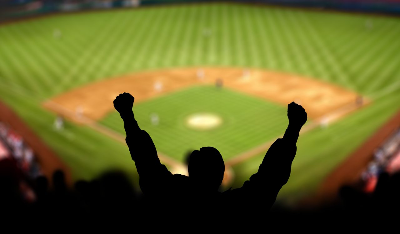 The silhouette of a baseball fan raising their arms in a baseball stadium.