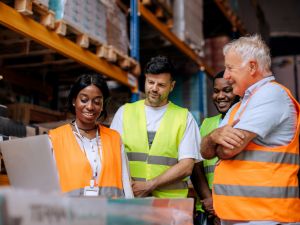 Four warehouse employees standing together in the aisle, focused on a laptop. They are reviewing inventory data while surrounded by shelves filled with boxes and packages.