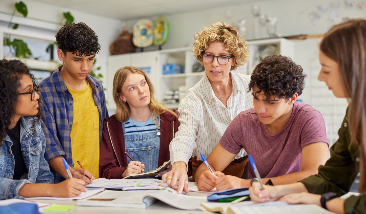 A group of teenagers working at a table listen to their teacher as she points out something in a book.