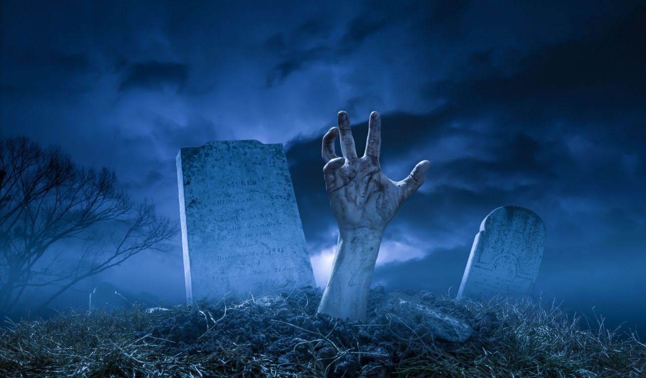 A zombie or monster hand pushing through the dirt at grave site in a graveyard on a dark Halloween night.