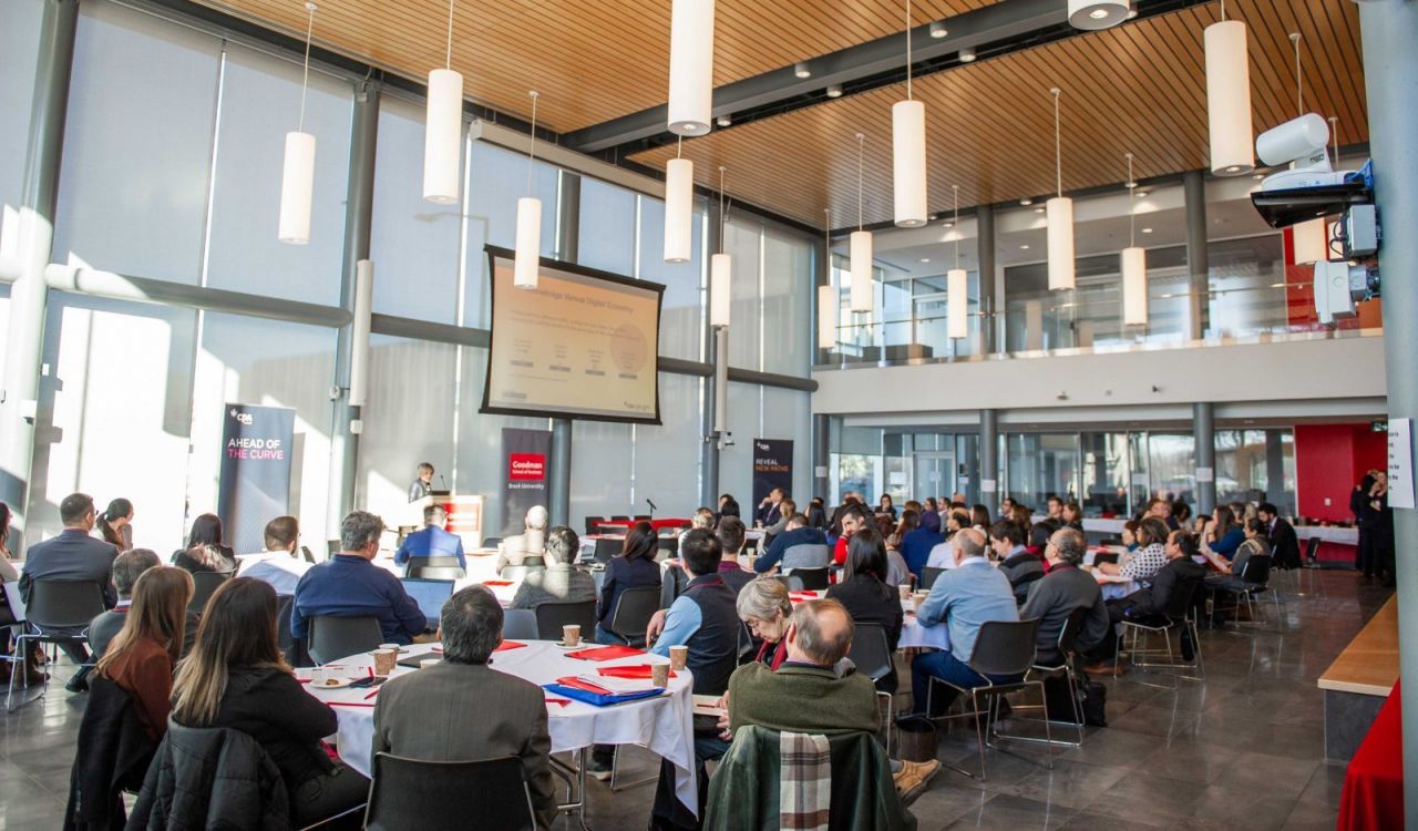 A room full of people at round tables look towards a podium at a symposium.