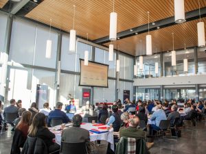 A room full of people at round tables look towards a podium at a symposium.