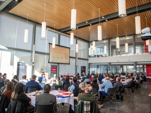 A room full of people at round tables look towards a podium at a symposium.