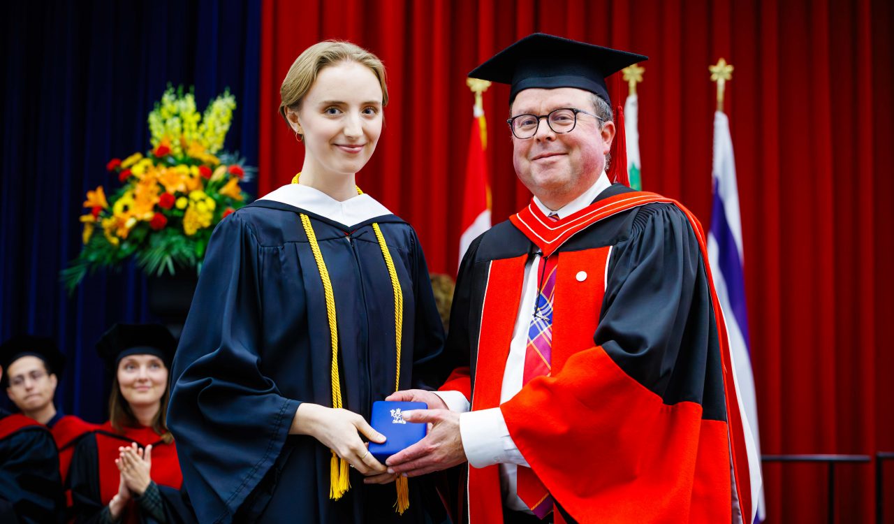 Two people in academic regalia pose with a medal on stage during a university graduation ceremony.