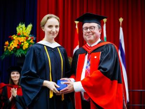 Two people in academic regalia pose with a medal on stage during a university graduation ceremony.