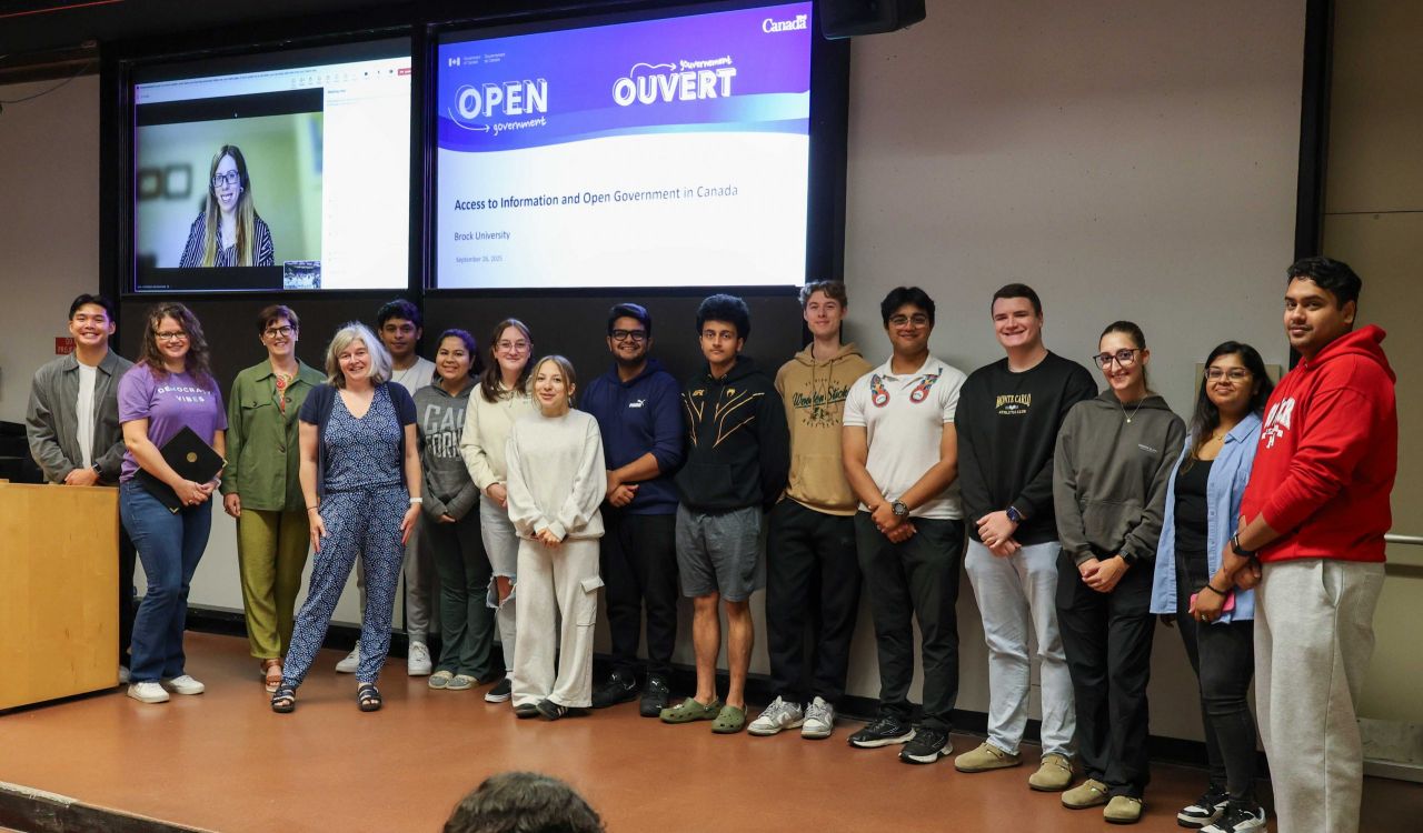 Ada Oliver (on screen), Junearn Lenon and Cara Scarfone of the Treasury Board of Canada Secretariat stand on stage with Brock’s Manager, Privacy & Records Management Marion Hansen, Associate Professor Karen Louise Smith and several COMM 3P19 students