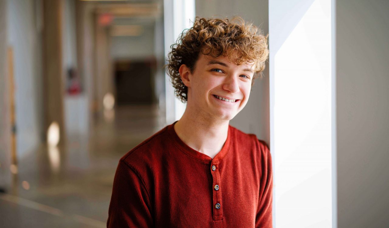 A young man stands in a brightly lit hallway with art on the on the wall.