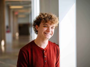 A young man stands in a brightly lit hallway with art on the on the wall.