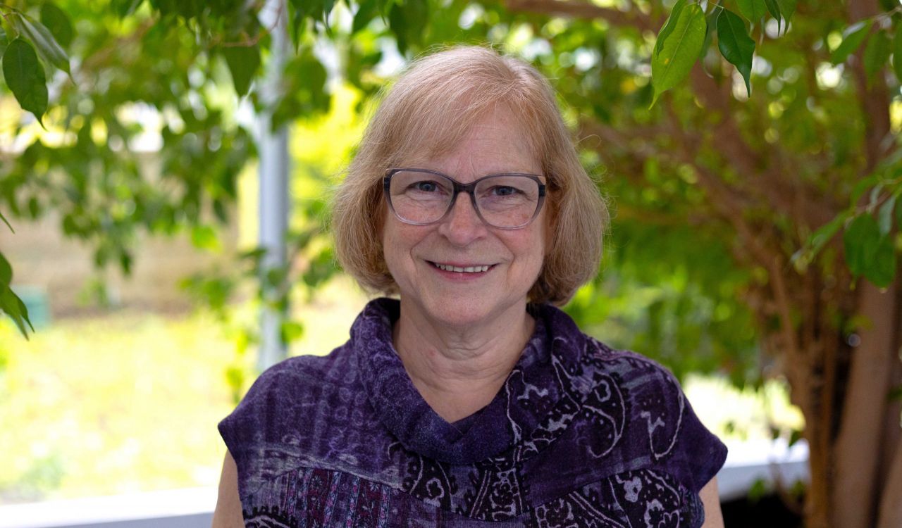 Woman stands in front of a tree with a garden in the background.