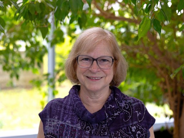 Woman stands in front of a tree with a garden in the background.