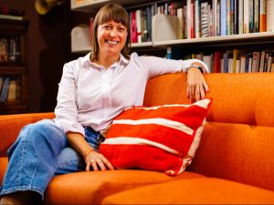 A woman wearing a white button-up shirt sits on an orange couch with an orange pillow in an office lined with books. 