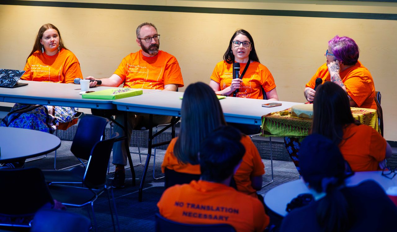 Four people sit together at a table in front of a group of other people.