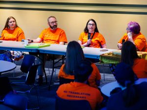 Four people sit together at a table in front of a group of other people.