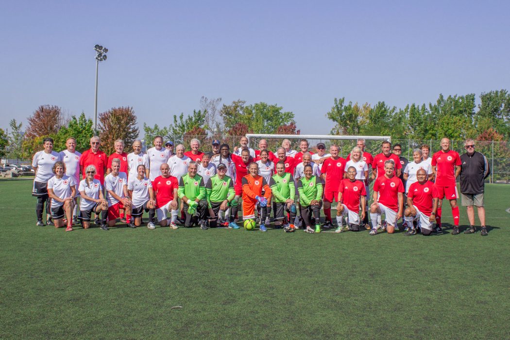 A large group of people in soccer uniforms pose for a group photo outside on a sunny day. 
