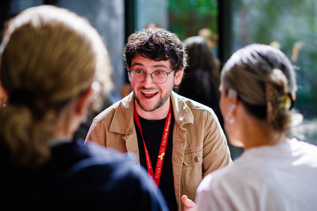 A young man wearing glasses and a tan jacket, smiling warmly and speaking animatedly with two people in the foreground