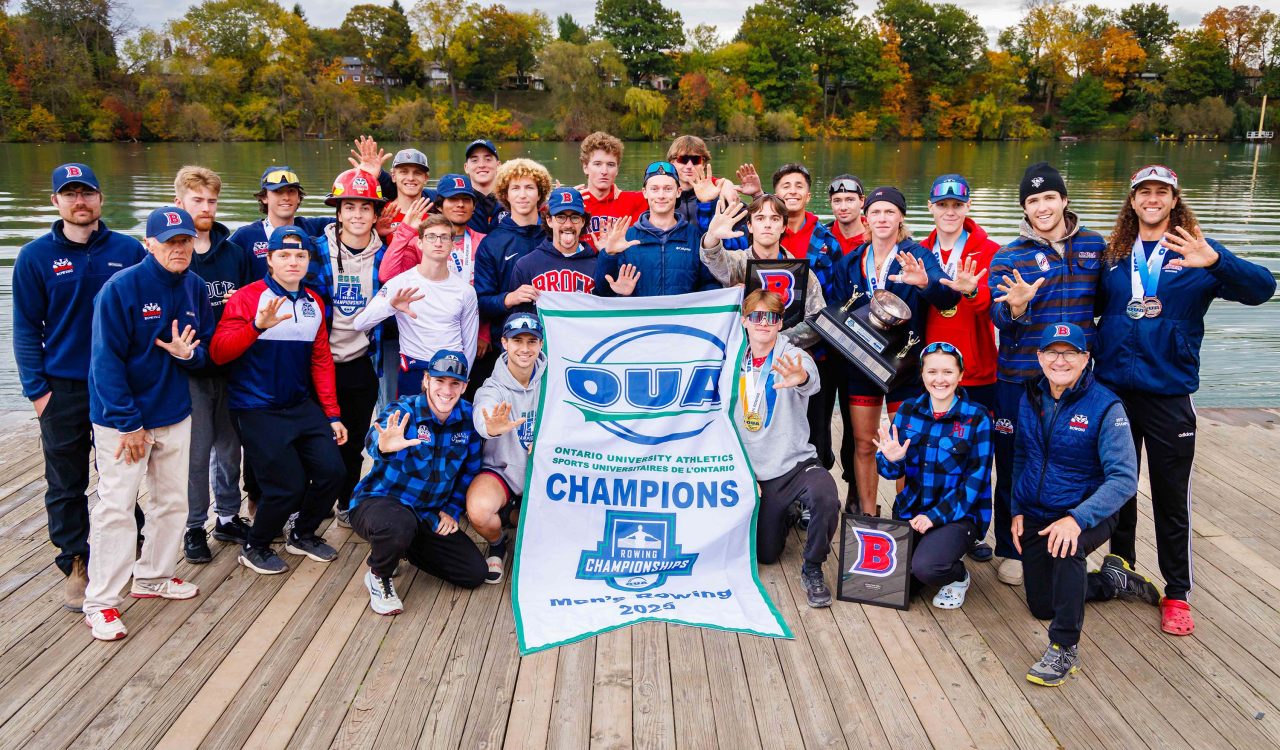 A group of men holding an OUA champions banner stand on a dock with a waterway behind them.