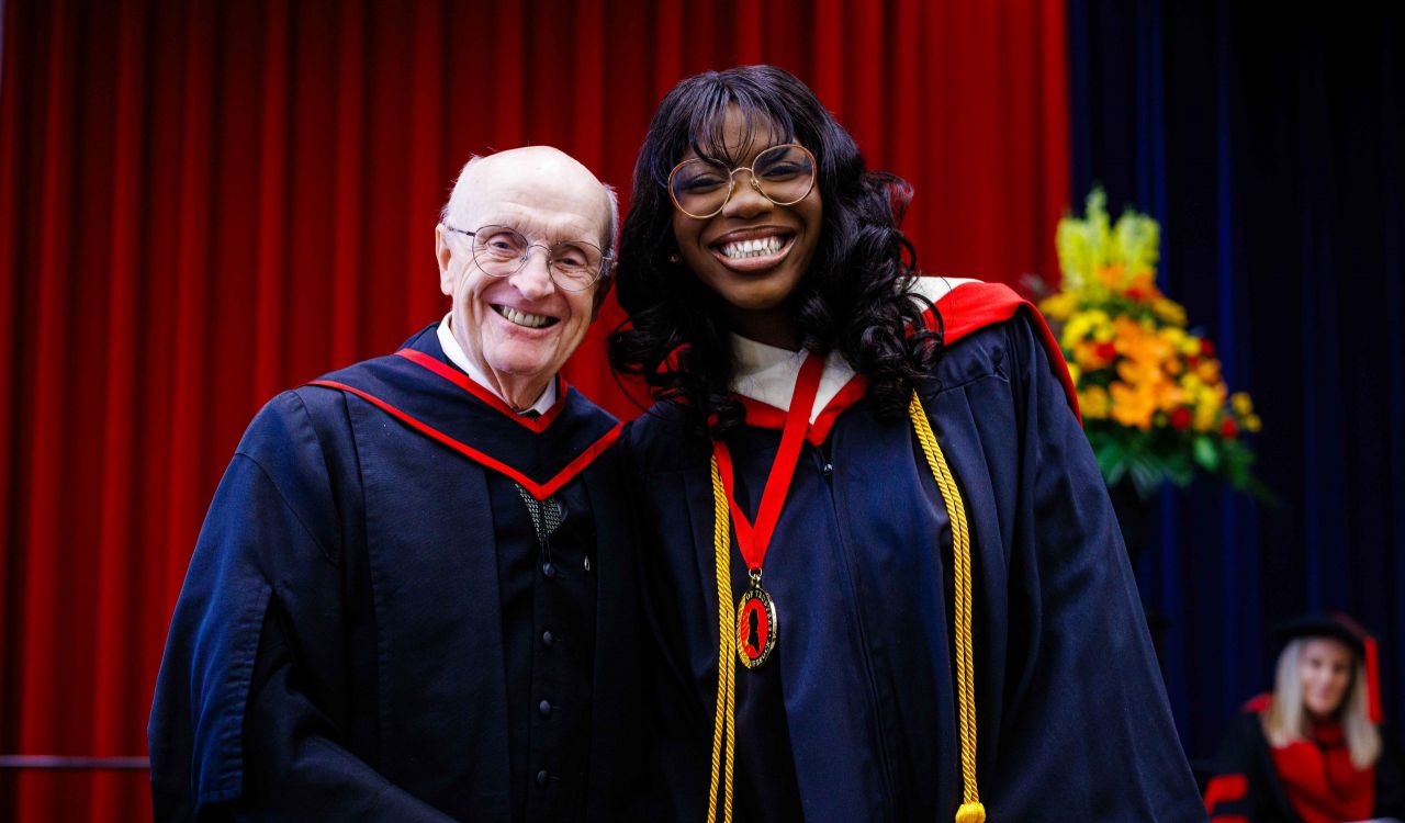 Two people in graduation gowns smile together on stage during a convocation ceremony. The person on the left is an older man wearing glasses, and the person on the right is a young woman wearing a medal and honor cords.
