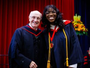 Two people in graduation gowns smile together on stage during a convocation ceremony. The person on the left is an older man wearing glasses, and the person on the right is a young woman wearing a medal and honor cords.