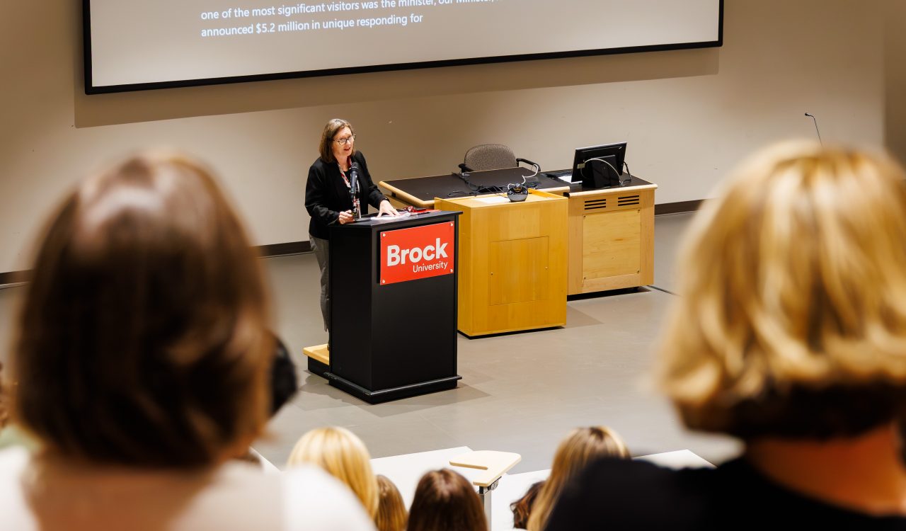 A woman stands at a podium speaking in an auditorium filled with people.
