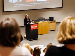 A woman stands at a podium speaking in an auditorium filled with people.