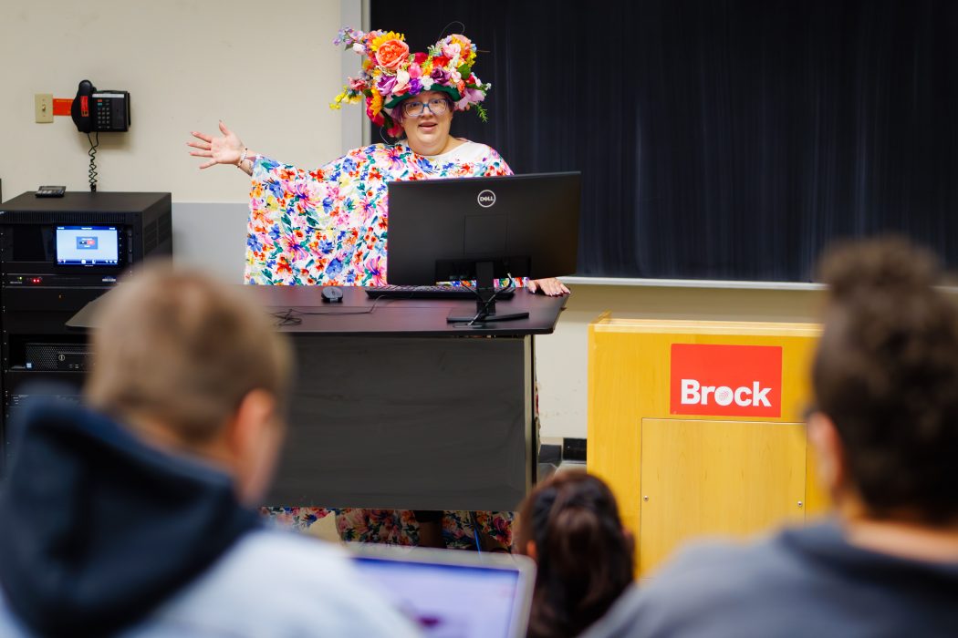 A woman in a floral dress and headdress stands behind a podium in a university classroom.
