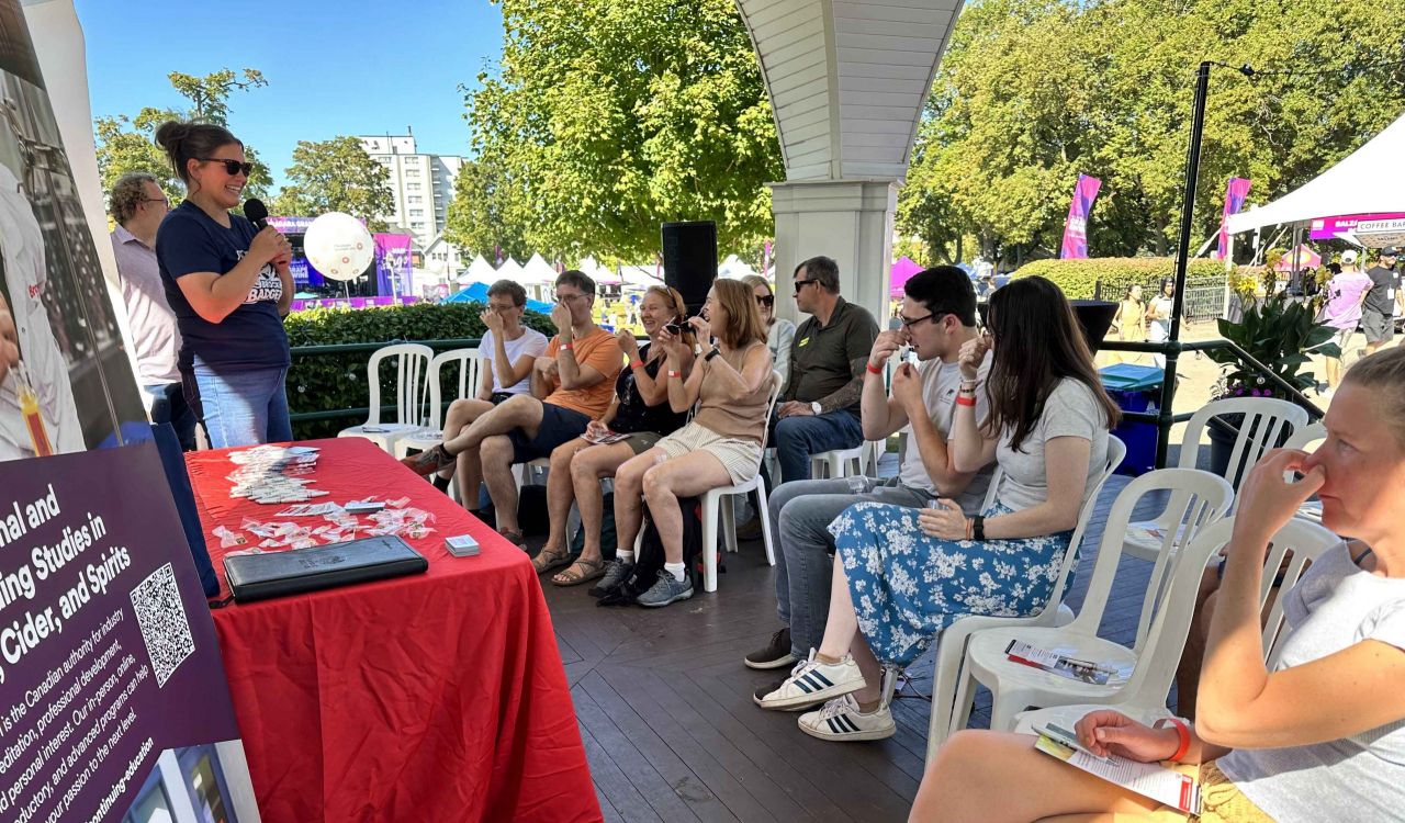 A group of people sit on chairs in rows in a covered outdoor space at a wine festival, plugging their noses as another person in front of them leads a wine sensory exercise.
