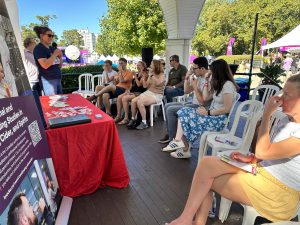 A group of people sit on chairs in rows in a covered outdoor space at a wine festival, plugging their noses as another person in front of them leads a wine sensory exercise.