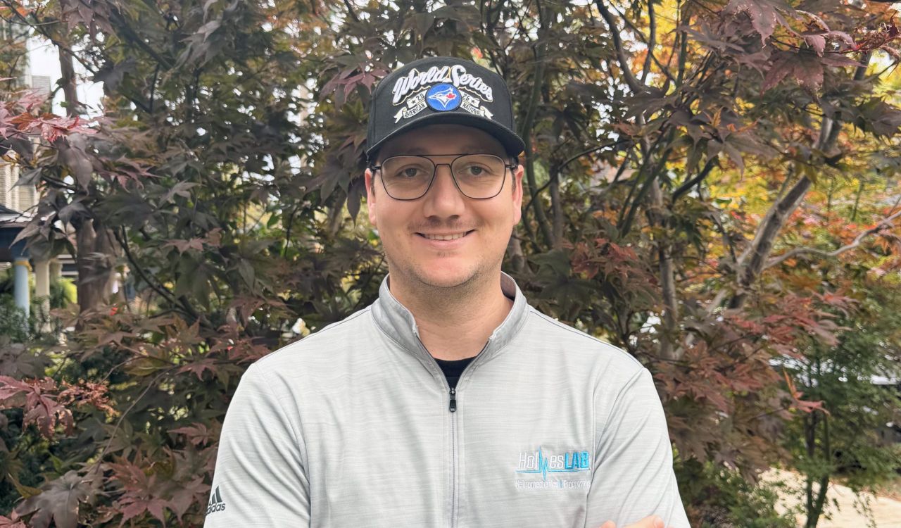 A man in a baseball cap poses outside in front of a garden.