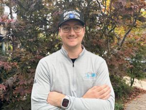 A man in a baseball cap poses outside in front of a garden.