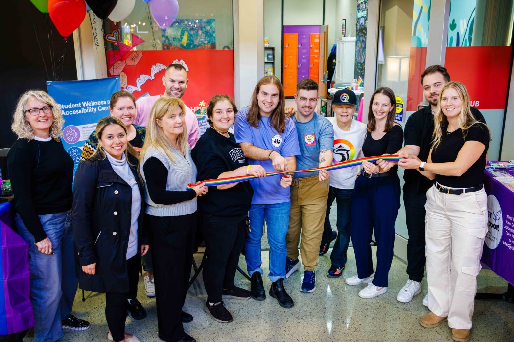A group of people smiling and standing in a semi-circle hold a rainbow ribbon for a ceremonial cutting inside a bright campus space decorated with balloons and posters.