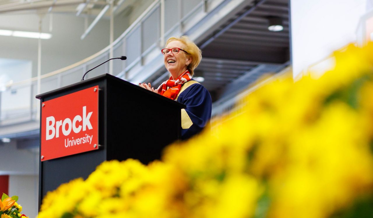 A woman stands at a podium with a red Brock University sign. Yellow flowers appear prominently in the foreground.