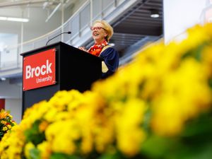 A woman stands at a podium with a red Brock University sign. Yellow flowers appear prominently in the foreground.