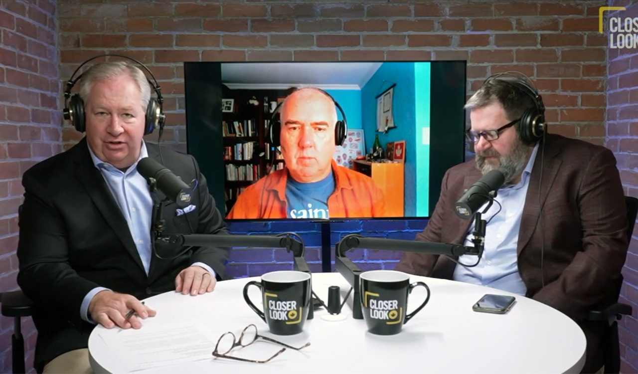 Two men at a desk talk to a man on a virtual screen during a podcast interview.