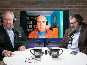 Two men at a desk talk to a man on a virtual screen during a podcast interview.
