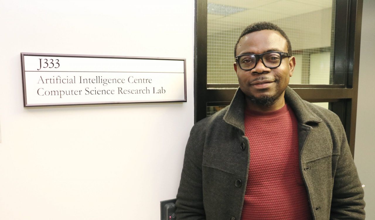 Assistant Professor of Computer Science Blessing Ogbuokiri stands next to a sign saying “J333. Artificial Intelligence Centre Computer Science Research Lab” with a window framing him in the background.