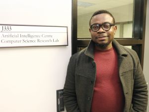 Assistant Professor of Computer Science Blessing Ogbuokiri stands next to a sign saying “J333. Artificial Intelligence Centre Computer Science Research Lab” with a window framing him in the background.