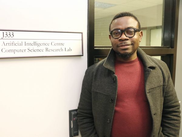Assistant Professor of Computer Science Blessing Ogbuokiri stands next to a sign saying “J333. Artificial Intelligence Centre Computer Science Research Lab” with a window framing him in the background.