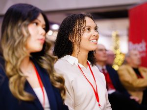 Two young women with red lanyards listen attentively during an event.