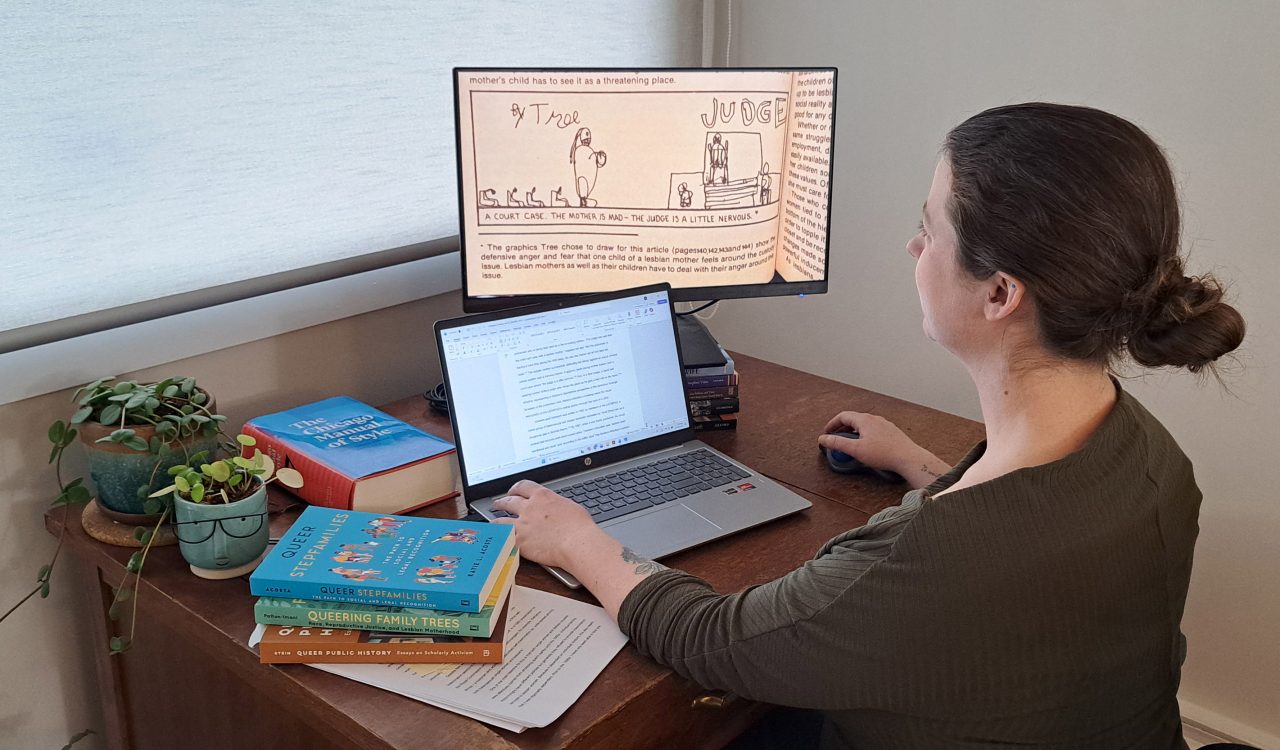 Profile of postdoctoral researcher Erin Gallagher-Cohoon sitting at a desk looking at two computer screens, one a laptop and other a larger screen depicting some drawings and words. Piles of books and two plans are also on the top of the desk.