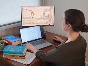 Profile of postdoctoral researcher Erin Gallagher-Cohoon sitting at a desk looking at two computer screens, one a laptop and other a larger screen depicting some drawings and words. Piles of books and two plans are also on the top of the desk.
