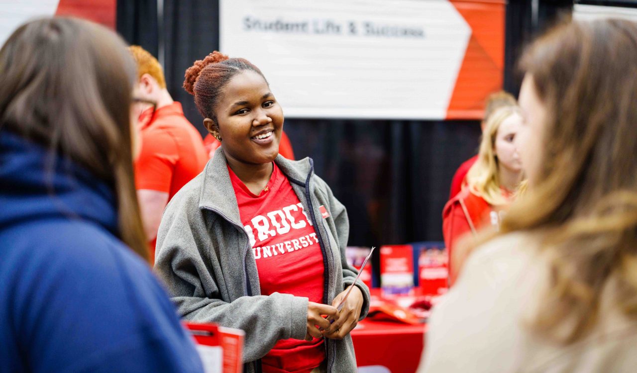 A smiling Brock University representative talks with visitors at an information booth during an event. The person is wearing a red Brock University shirt and grey jacket, standing in front of a red and white display labeled "Student Life and Success."