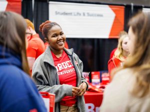 A smiling Brock University representative talks with visitors at an information booth during an event. The person is wearing a red Brock University shirt and grey jacket, standing in front of a red and white display labeled "Student Life and Success."