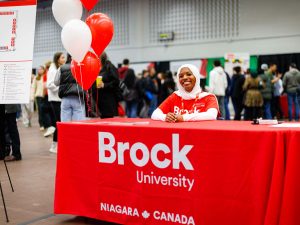 A smiling Brock University ambassador welcomes visitors at an information table decorated with red and white balloons.