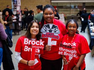 Three Brock student volunteers in red shirts pose with Brock University signs.