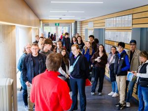 A tour guide in a red jacket speaks to a group of students and parents lined up in a hallway.