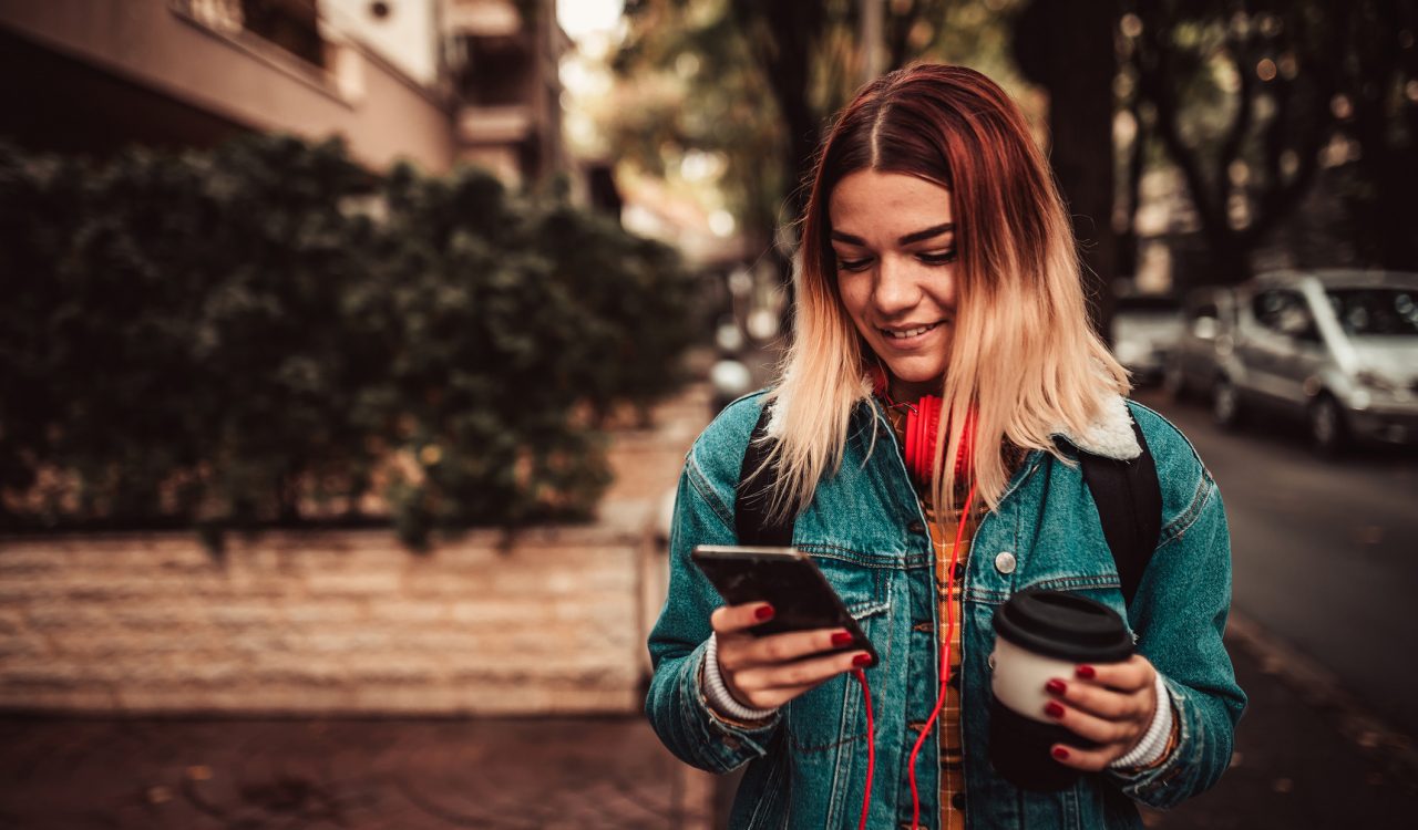 A teenage girl walks down a street while holding her cellphone and a coffee.