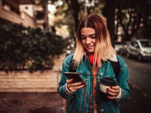 A teenage girl walks down a street while holding her cellphone and a coffee.