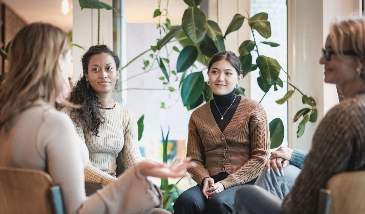 Four women sit in a circle having a conversation with a window and plant behind them.