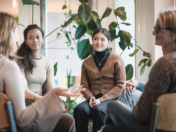 Four women sit in a circle having a conversation with a window and plant behind them.