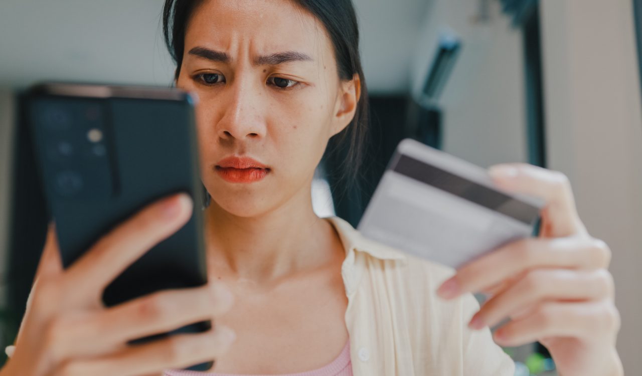 Close-up of a young woman who is looking at her phone with a concerned expression while holding a bank card.